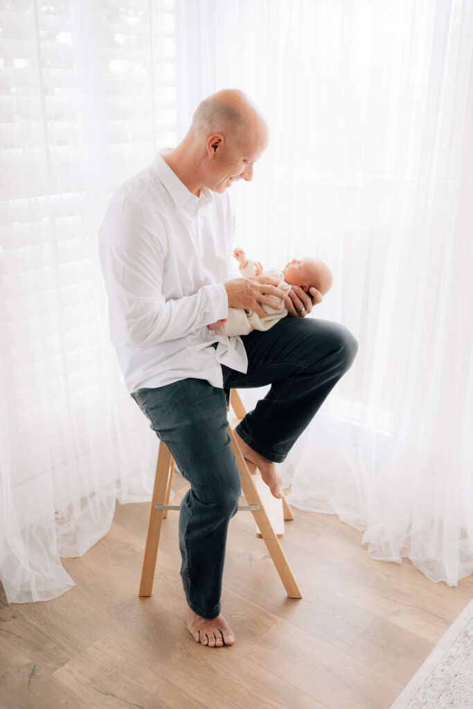 Denver photographer studio with dad sitting on stool holding newborn surrounded by bright windows 
