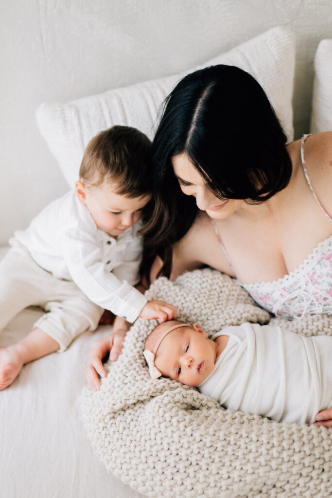 newborn baby lying on bed with mom and big brother who is reaching for her head