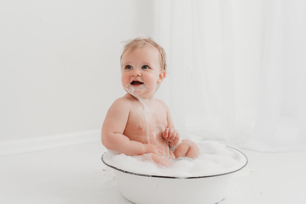 Baby is splashing in rustic basin tub for her first birthday studio milestone session after her cake smash