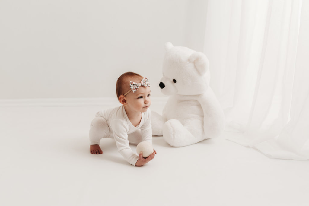 baby holding a white ball and crawling away from a stuffed bear during her first birthday studio milestone session