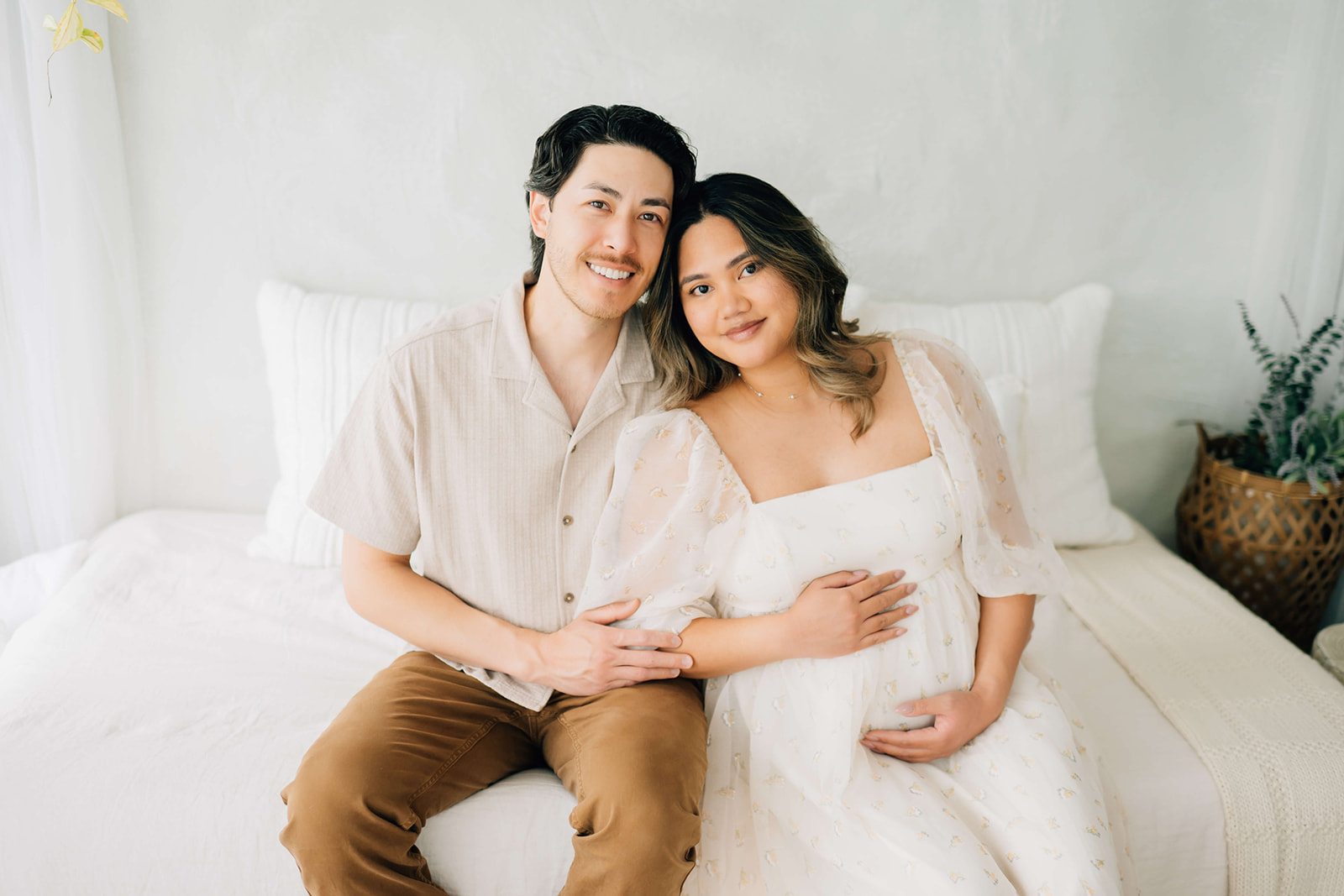 An expecting couple sits on a bed with hands on the bump and big smiles in a studio