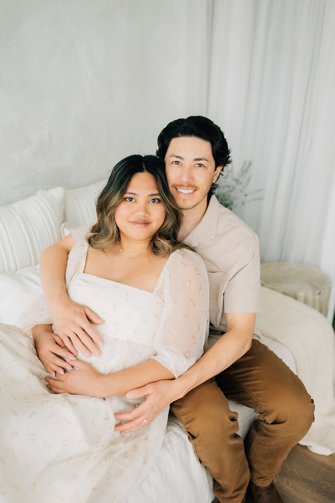 A happy expecting couple in white and tan snuggle on a bed in a studio after getting a 3D ultrasound in Denver
