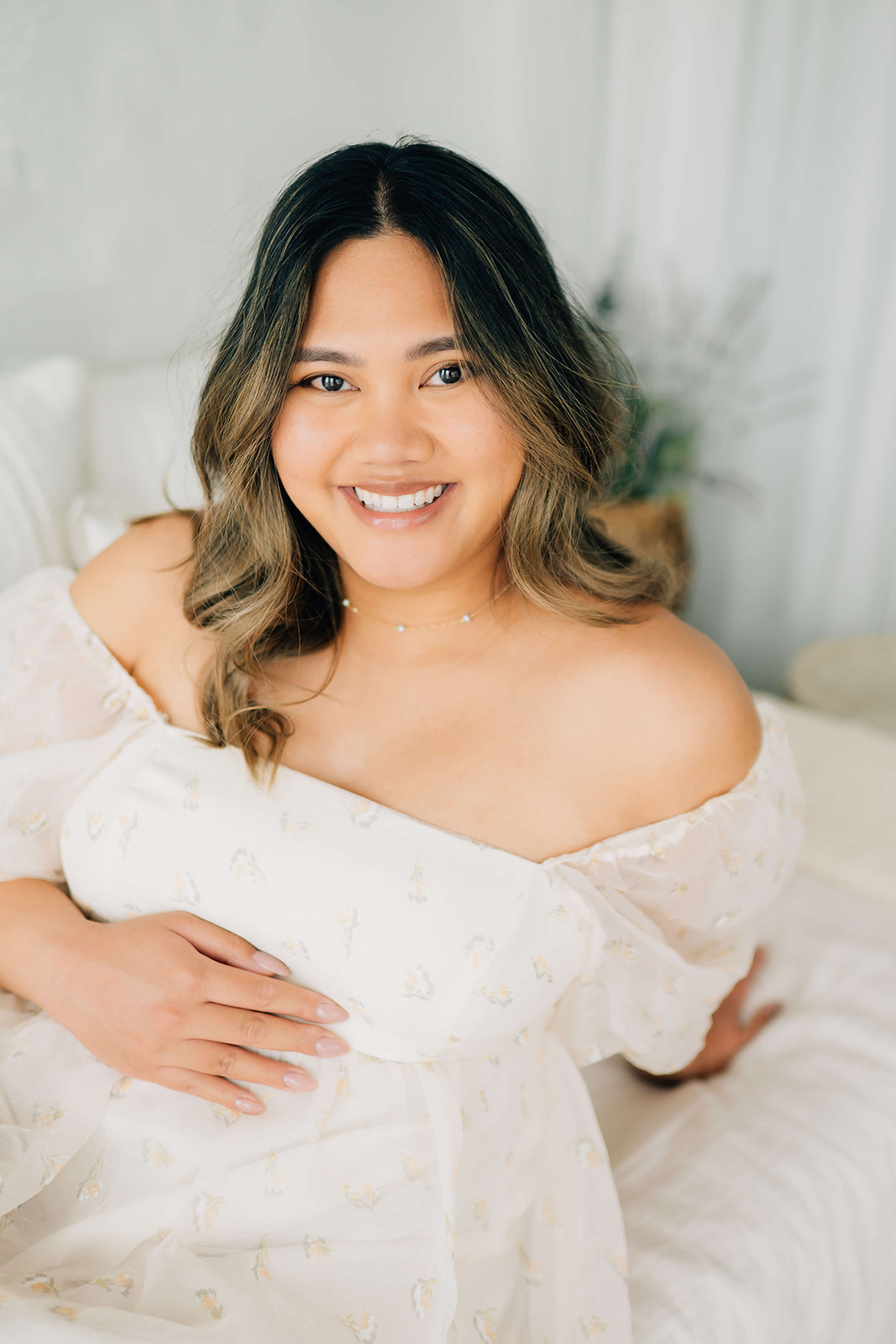 A happy mom to be in a white off the shoulder gown leans back on a bed smiling after a 3D ultrasound in Denver