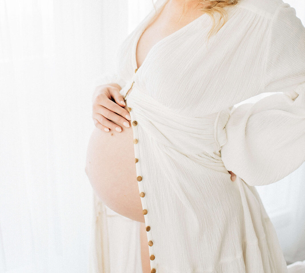 A smiling mother to be in a white open gown looks down to her hand on the bump while standing in a window