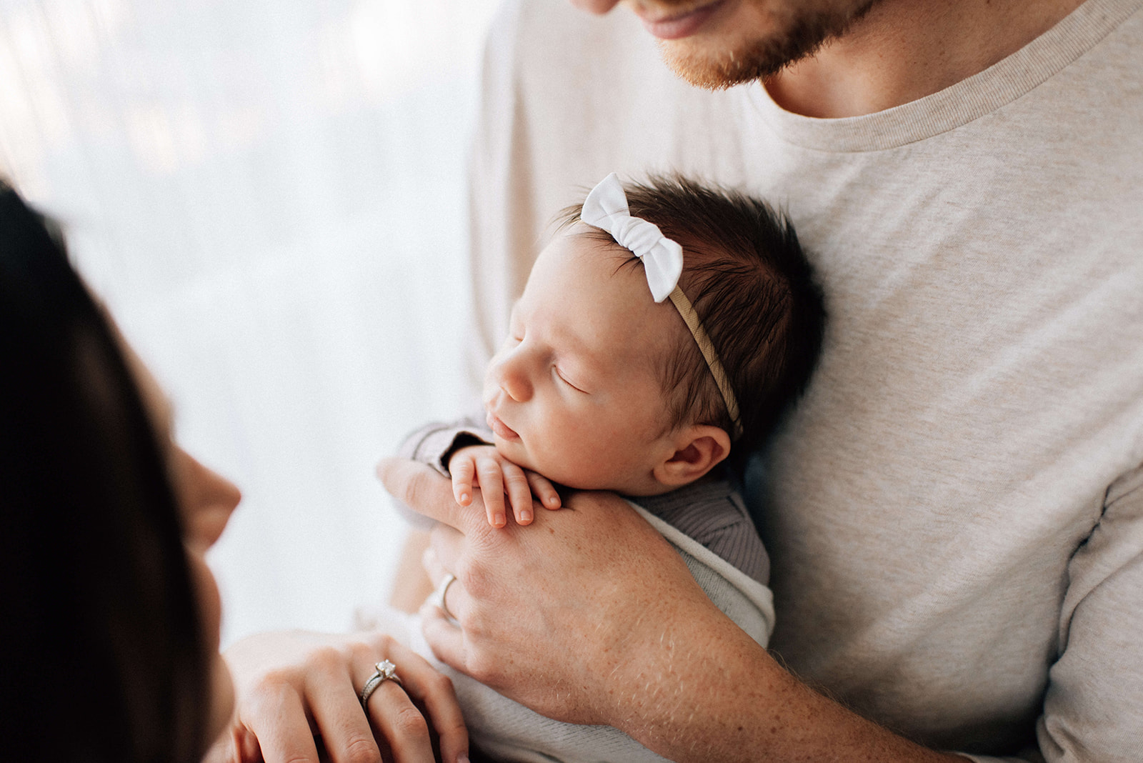A newborn baby girl sleeps against dad's chest with a white bow headband after visiting baby stores in Denver