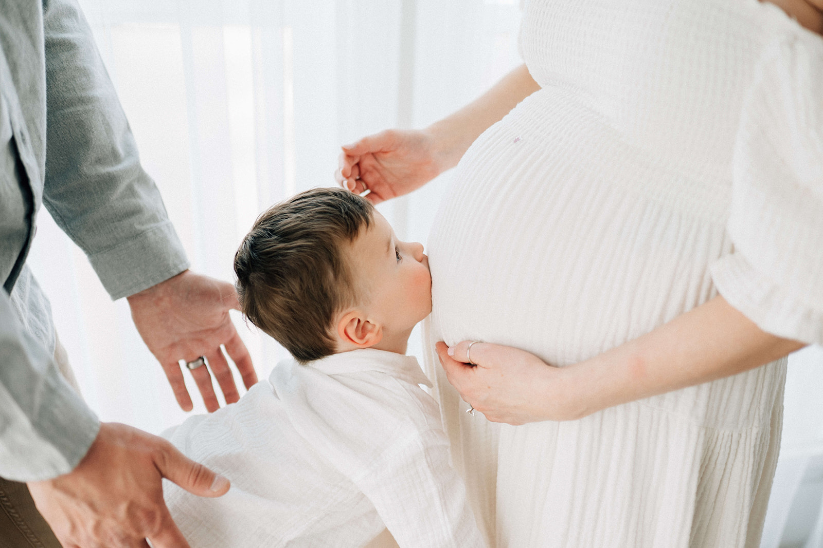 A toddler kisses mom's pregnant belly in a white shirt and white maternity dress