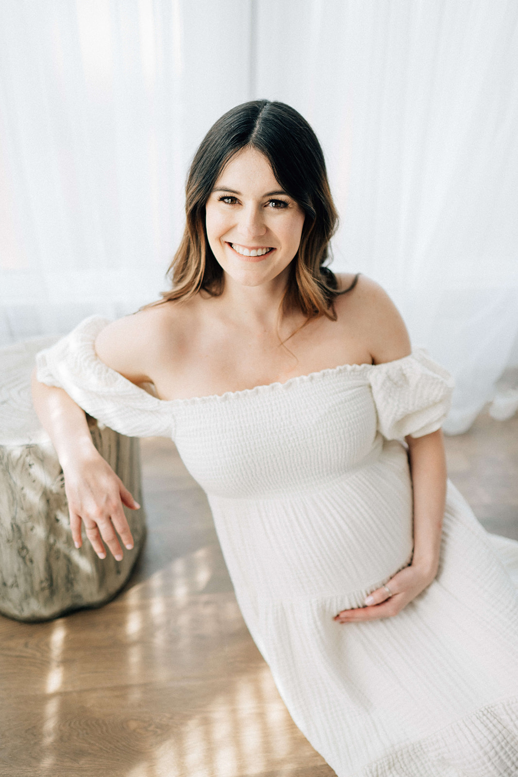 A smiling mother to be leans on a stool on the floor of a studio in a white maternity gown after visiting birthing centers in Denver
