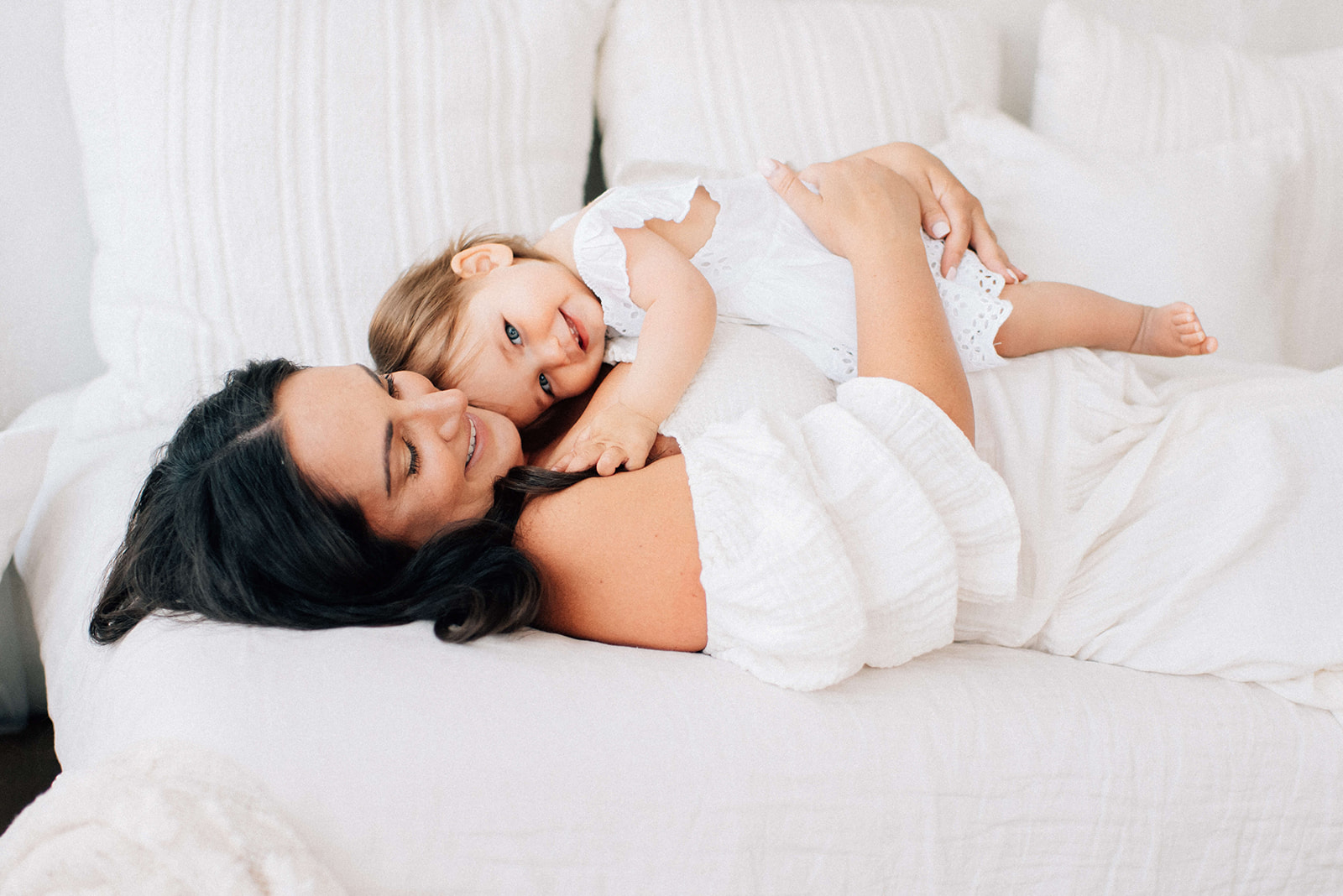 A giggling toddler girl snuggles on mom's chest while they lay on a bed in white dresses