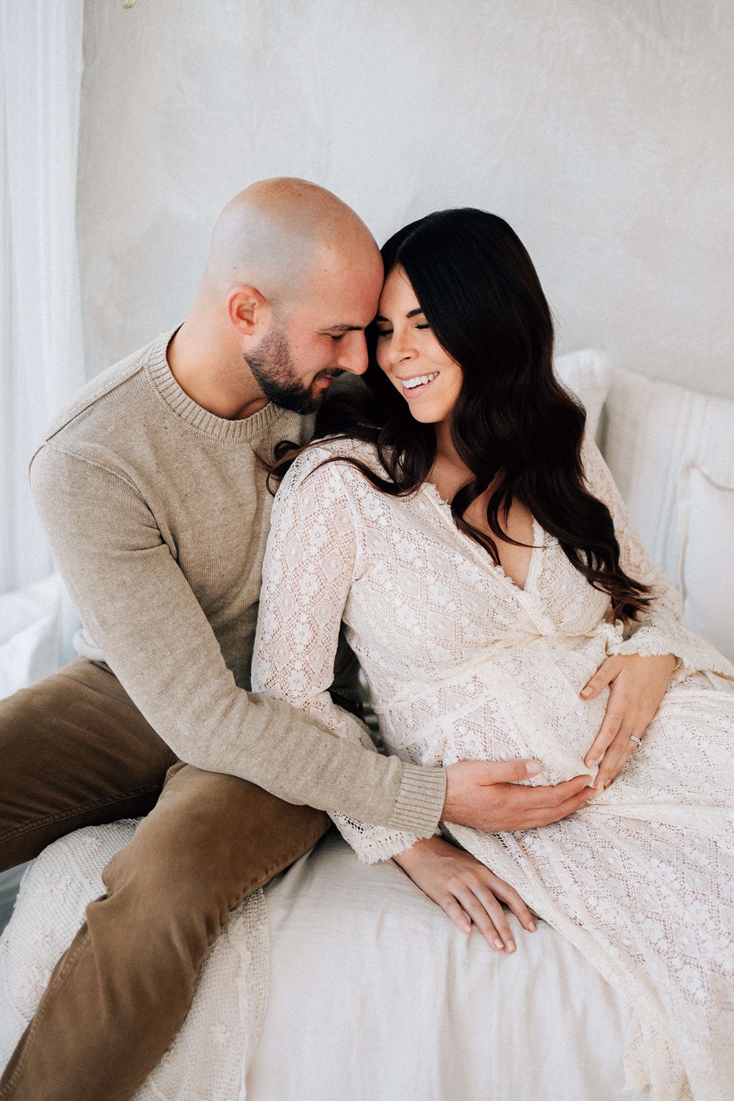 A pregnant couple snuggles on the bed with big smiles and hands on the bump in a sweater and lace gown after meeting Denver Doulas