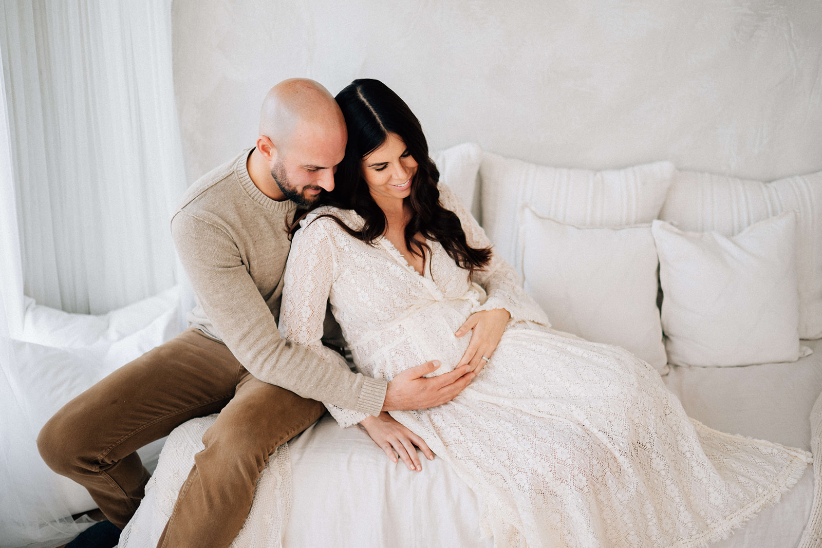 A happy expecting couple sits on a bed with hands on the bump after meeting some Denver Doulas