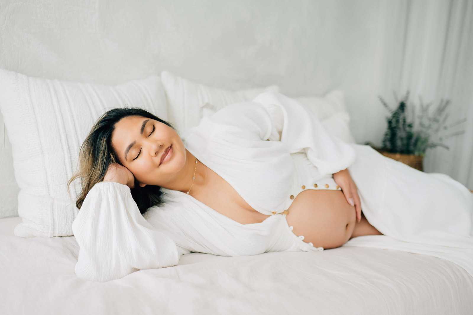 A glowing mother to be lays across a bed in an open white maternity gown