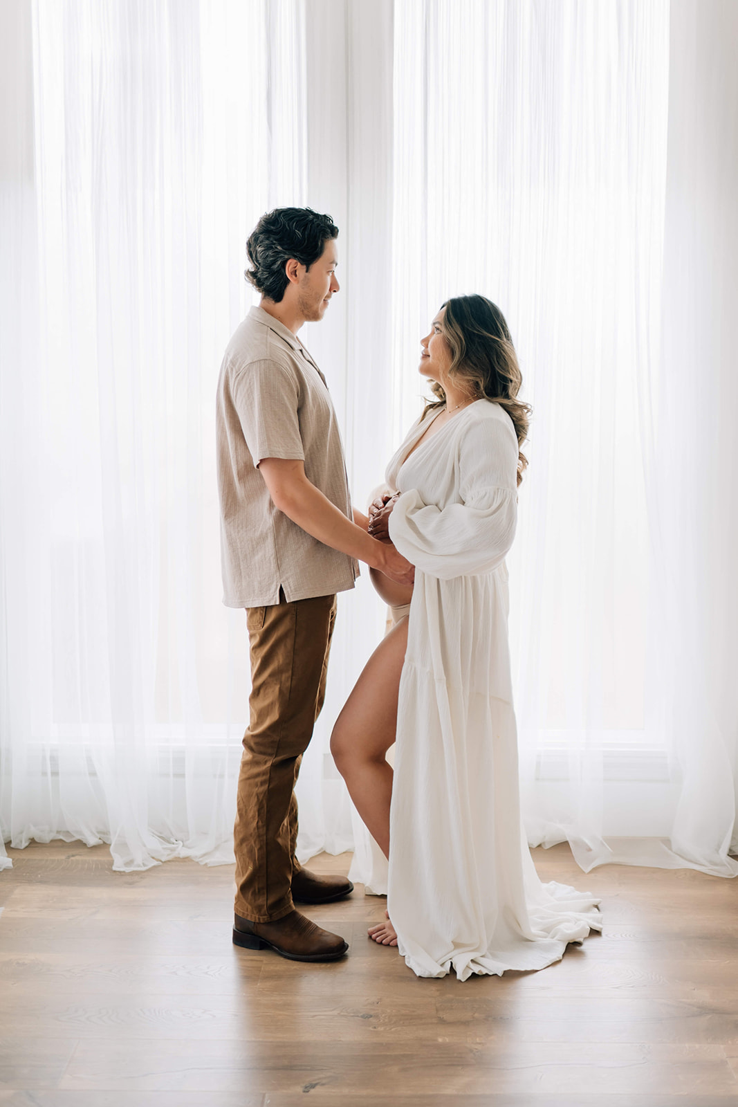 A happy expecting couple stands in a tall window of a studio holding the bump and smiling at each other after finding Denver midwives