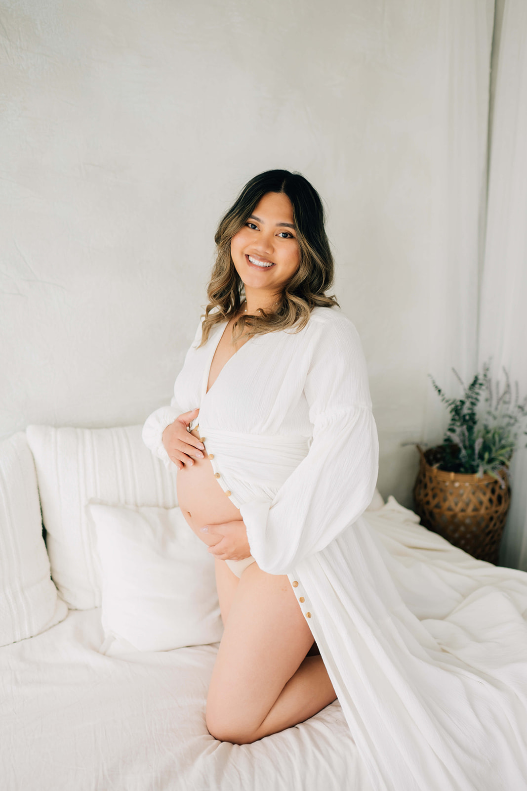 A happy mom to be kneels on a bed holding her bump in an unbuttoned white dress after finding Denver midwives