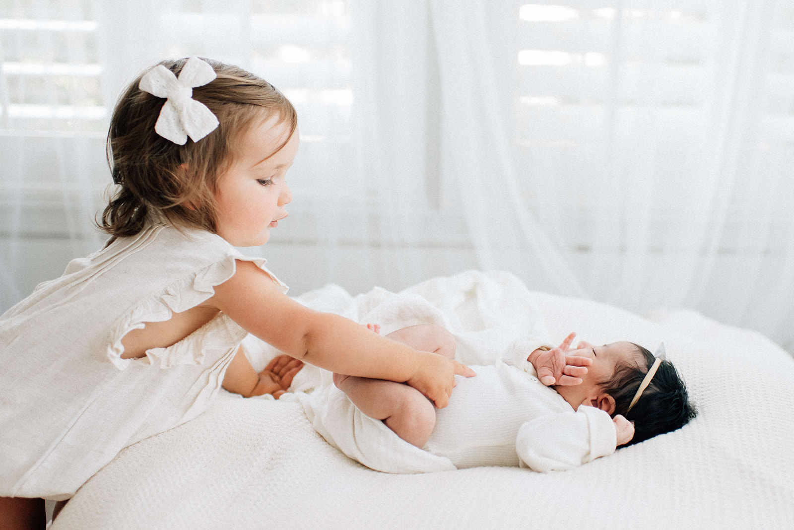A toddler girl in a white dress reaches to touch her sleeping newborn baby sister on a bed