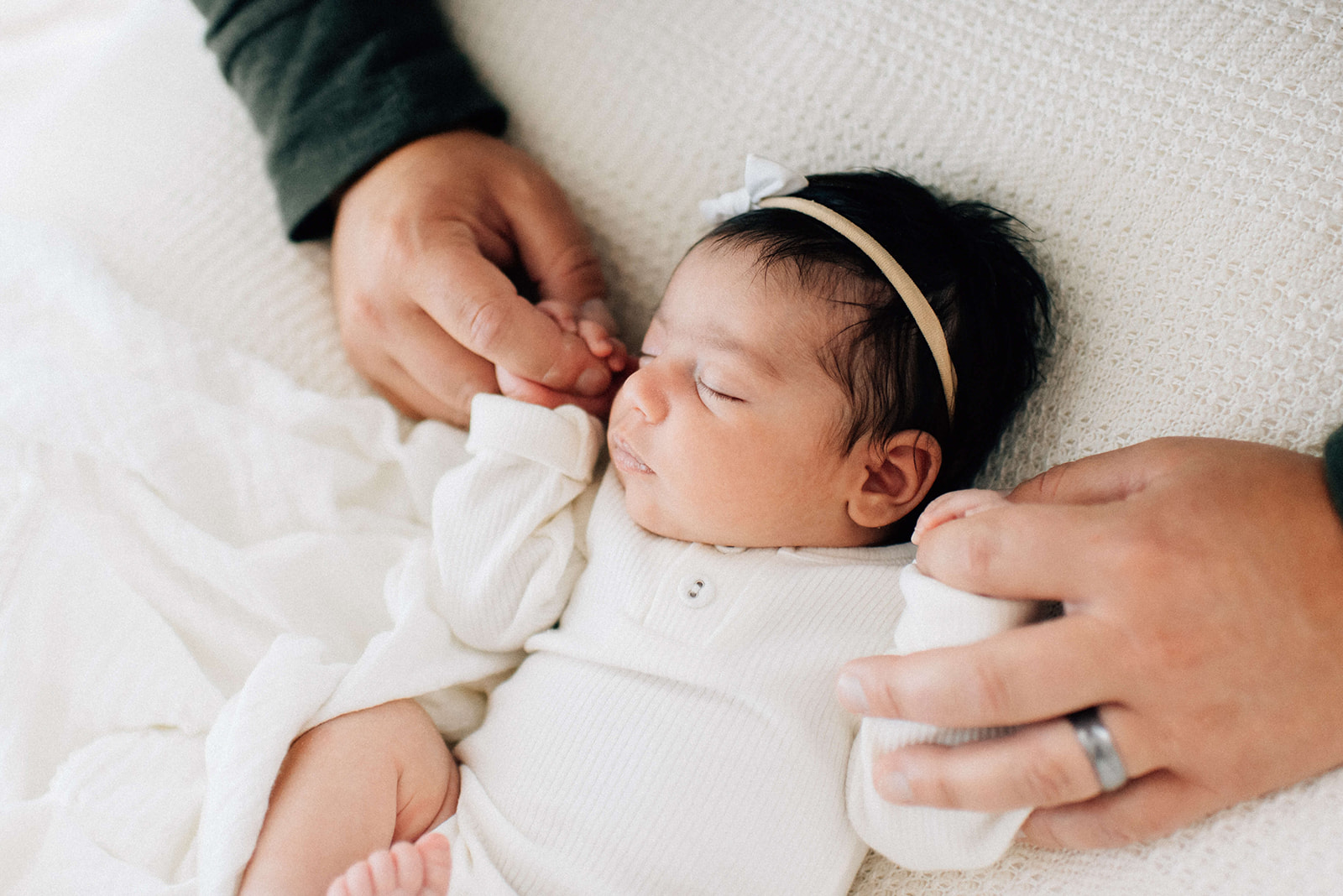 A newborn baby girl sleeps in a white onesie while holding dad's fingers in both hands while susing a diaper service in Denver