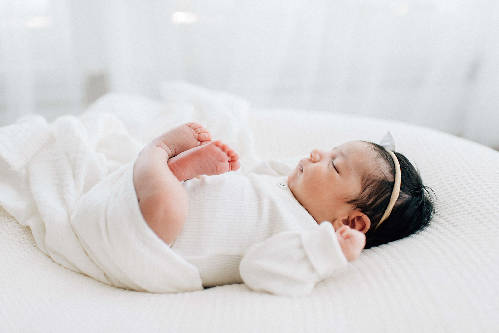 A newborn baby girl with dark hair sleeps on her back on a white bed in a matching onesie while using a diaper service in Denver