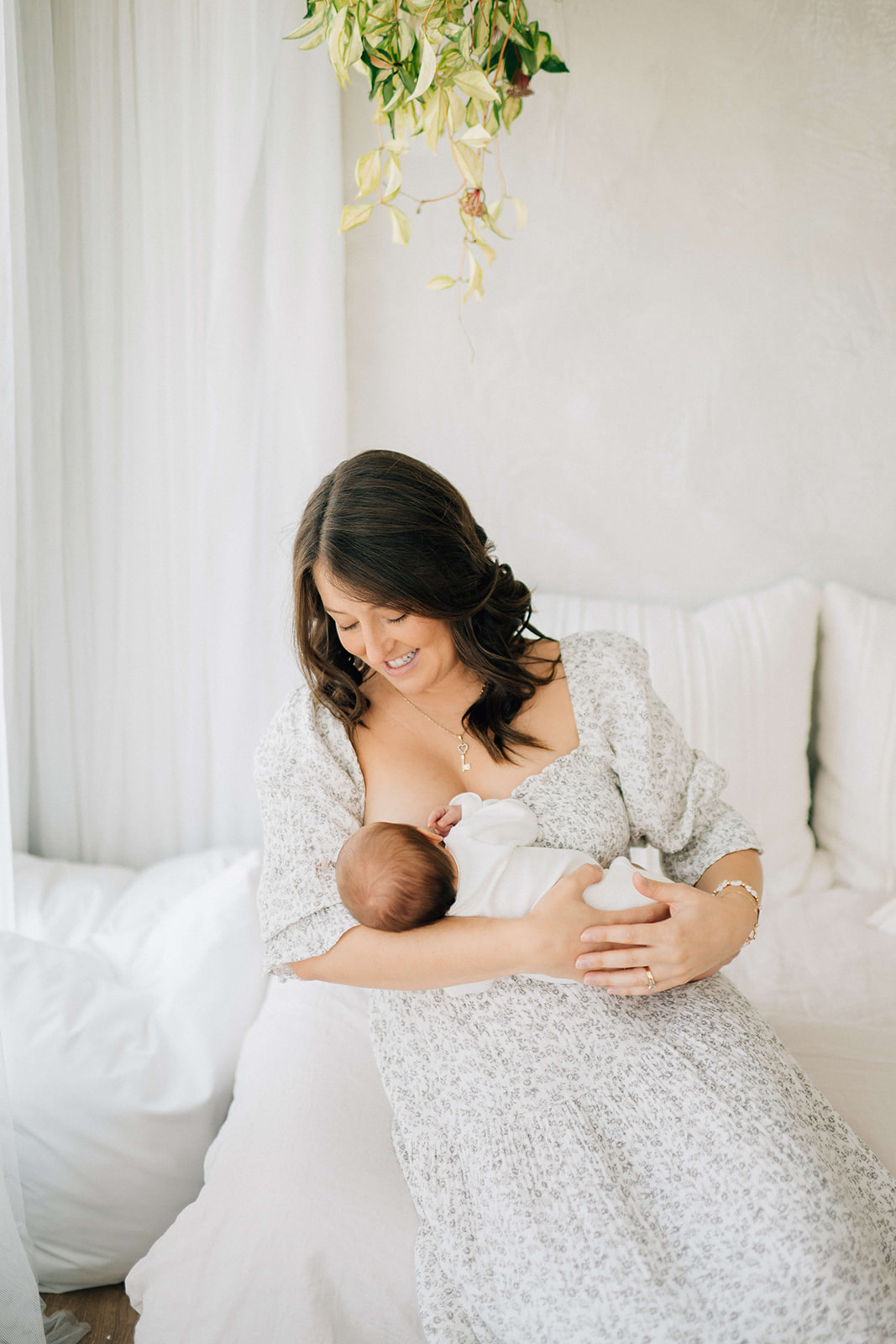 A smiling new mom in a white floral dress breastfeeds her newborn baby while sitting on a bed