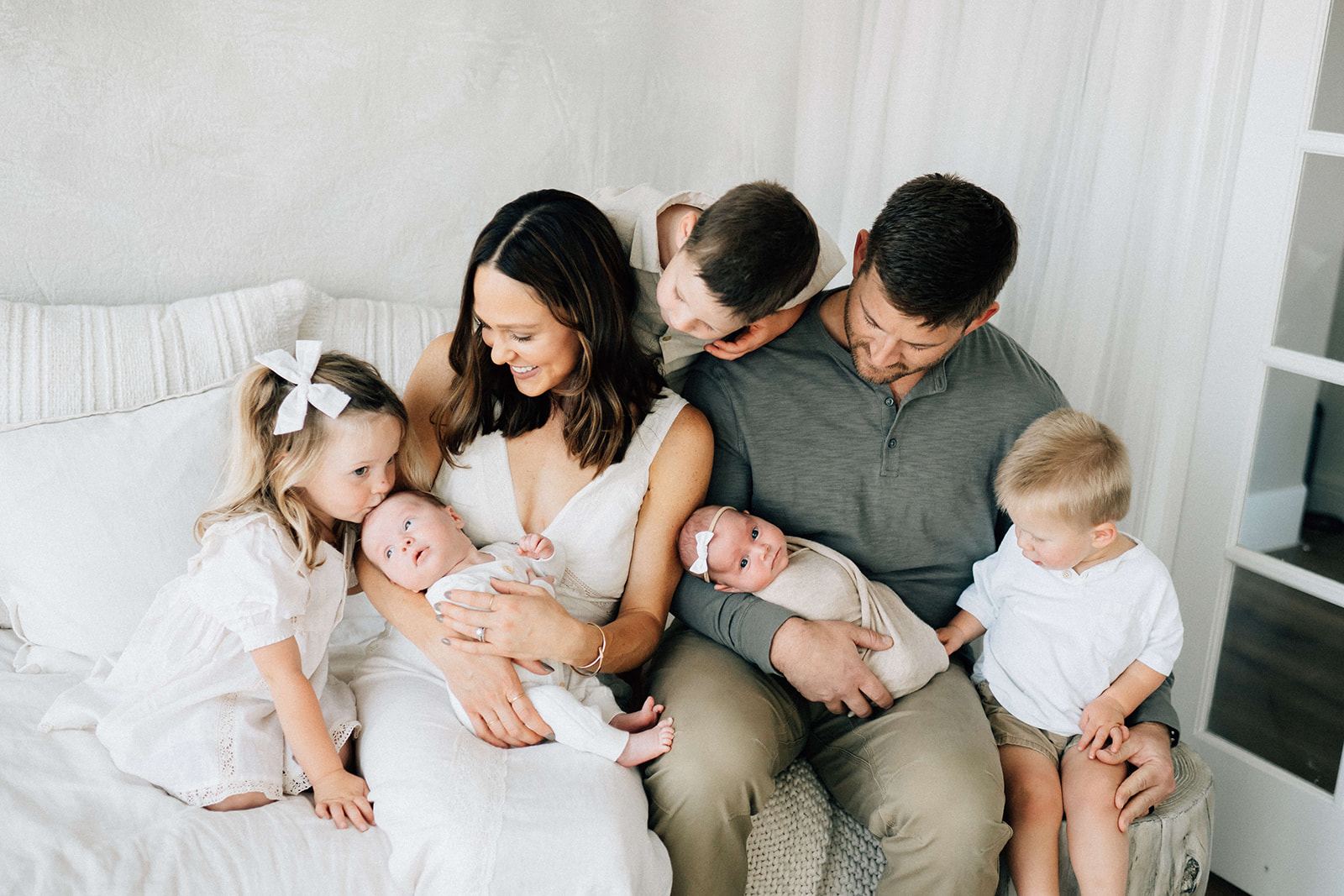 Three toddler siblings kiss and peek at their twin newborn siblings in mom and dad's lap on a couch