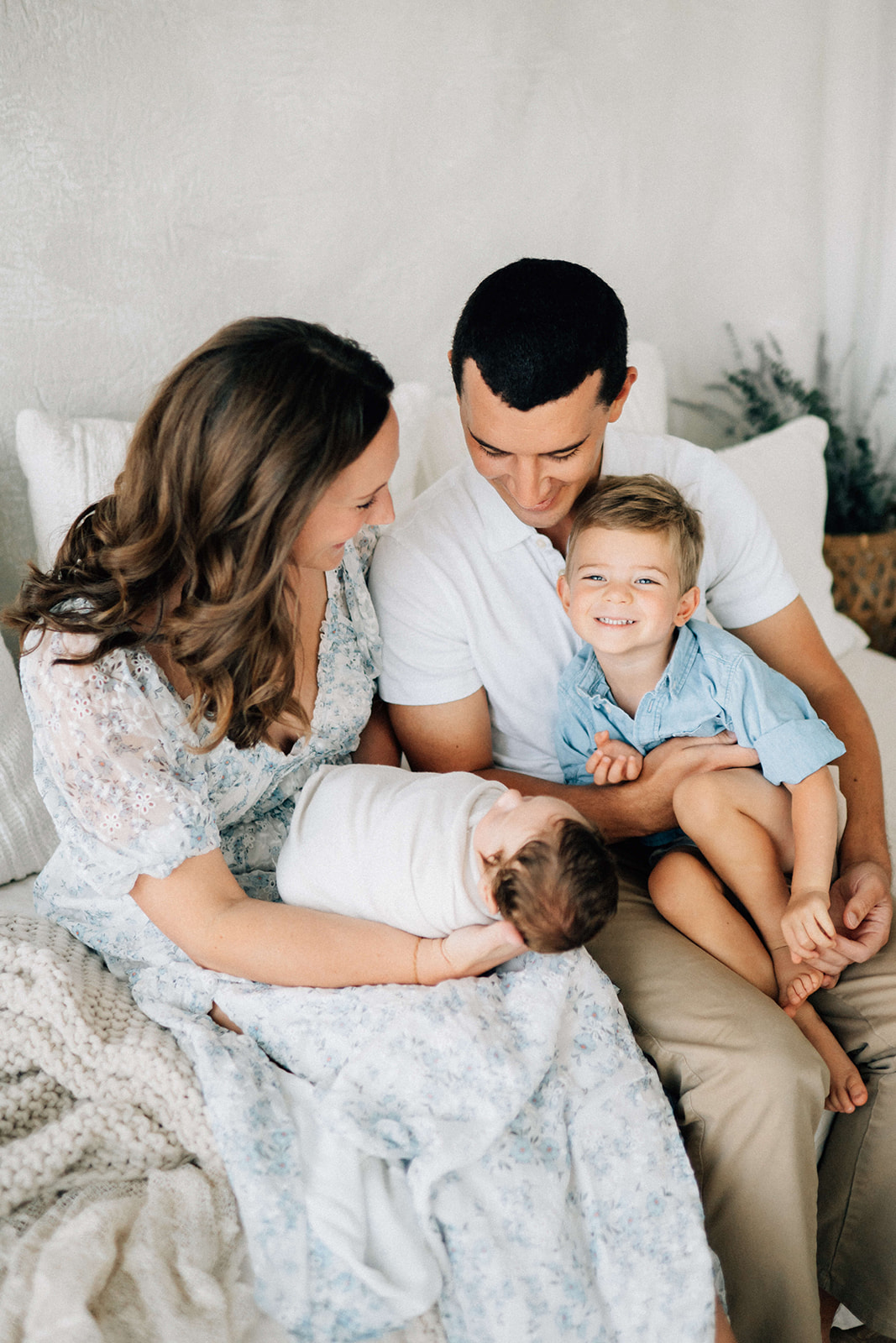 A happy toddler sits in dad's lap on a bed with mom holding a newborn after visiting pediatric dentist in Denver