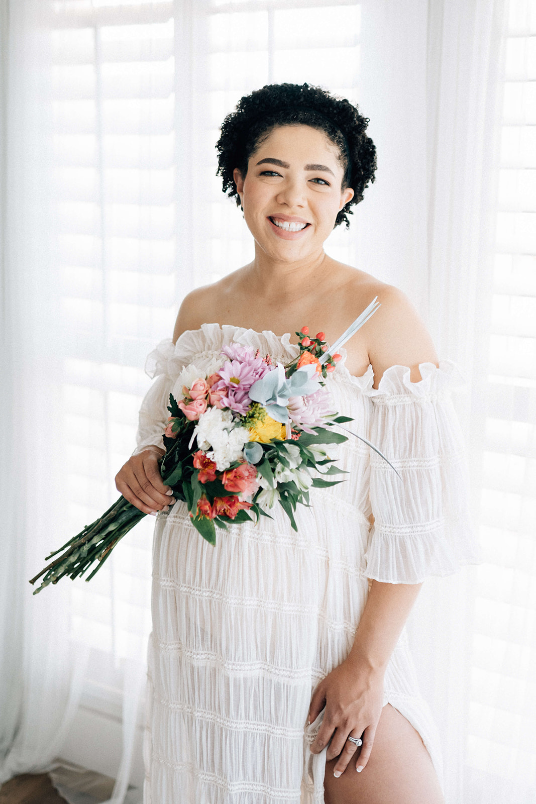 A happy expecting woman stands smiling in a white maternity gown and bright bouquet in a studio