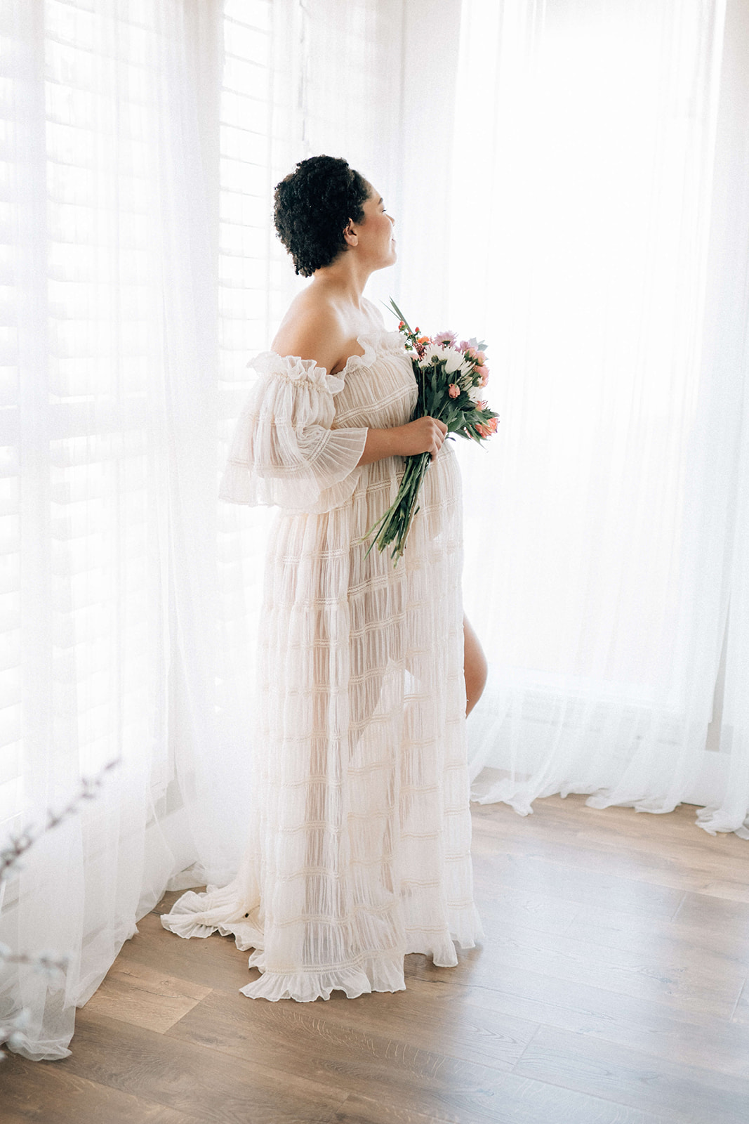 A pregnant woman in a white gown smiles into a window while standing with a colorful bouquet after some pelvic floor therapy in Denver