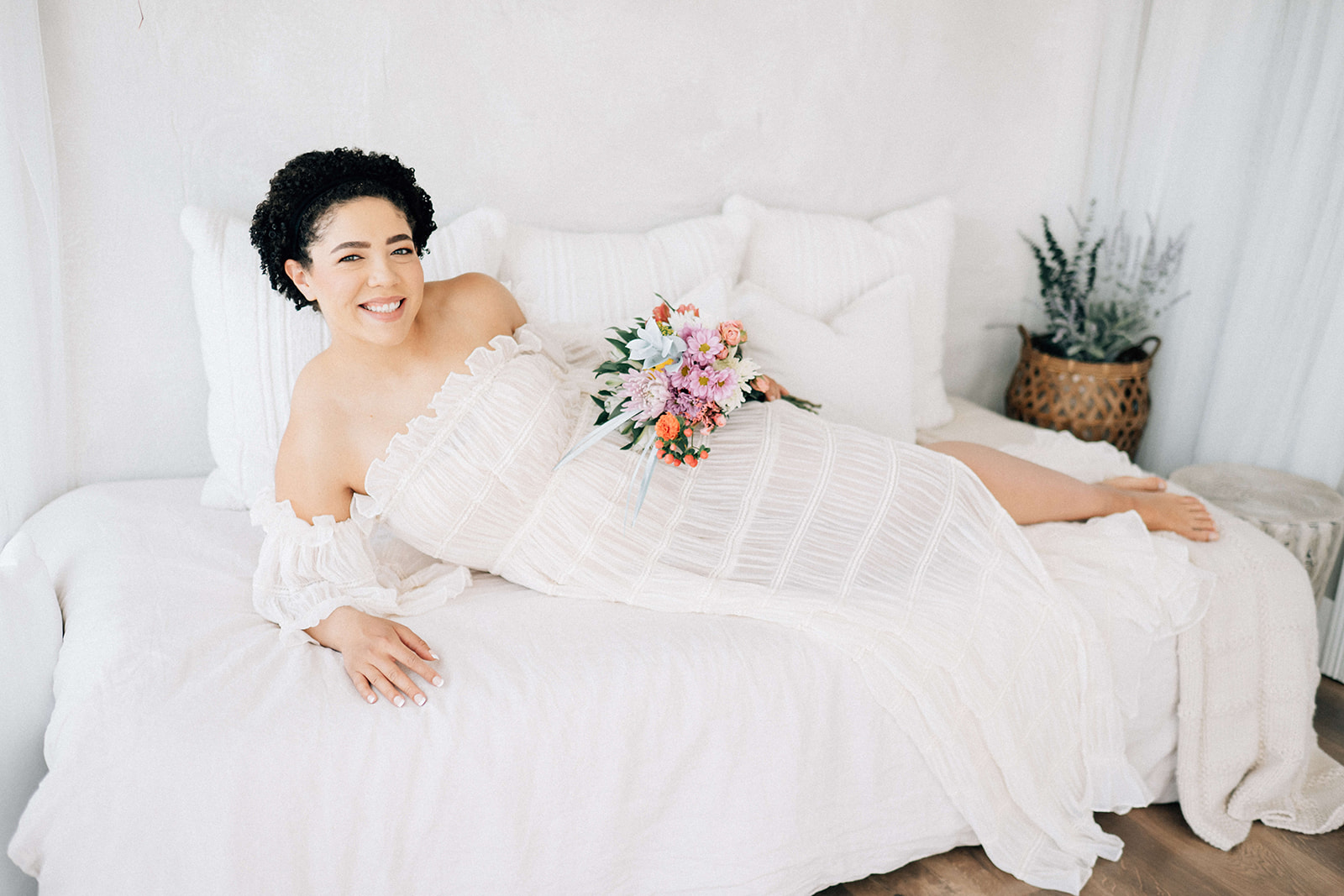 A smiling mother to be lays across a bed holding a colorful bouquet in a white gown after some pelvic floor therapy in Denver