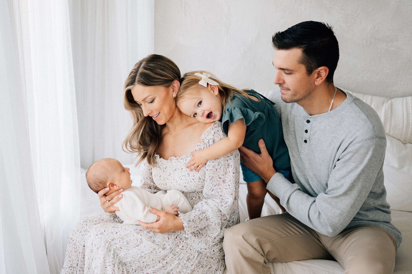 A toddler girl hugs mom while standing on a bed with mom and dad admiring their newborn in mom's lap