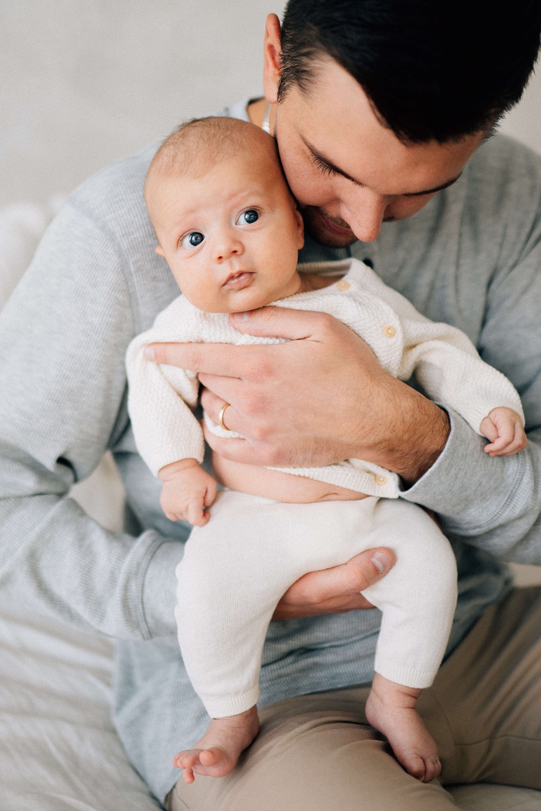 A baby sits in dad's hands while on a bed in a white onesie after using postpartum doulas in Denver