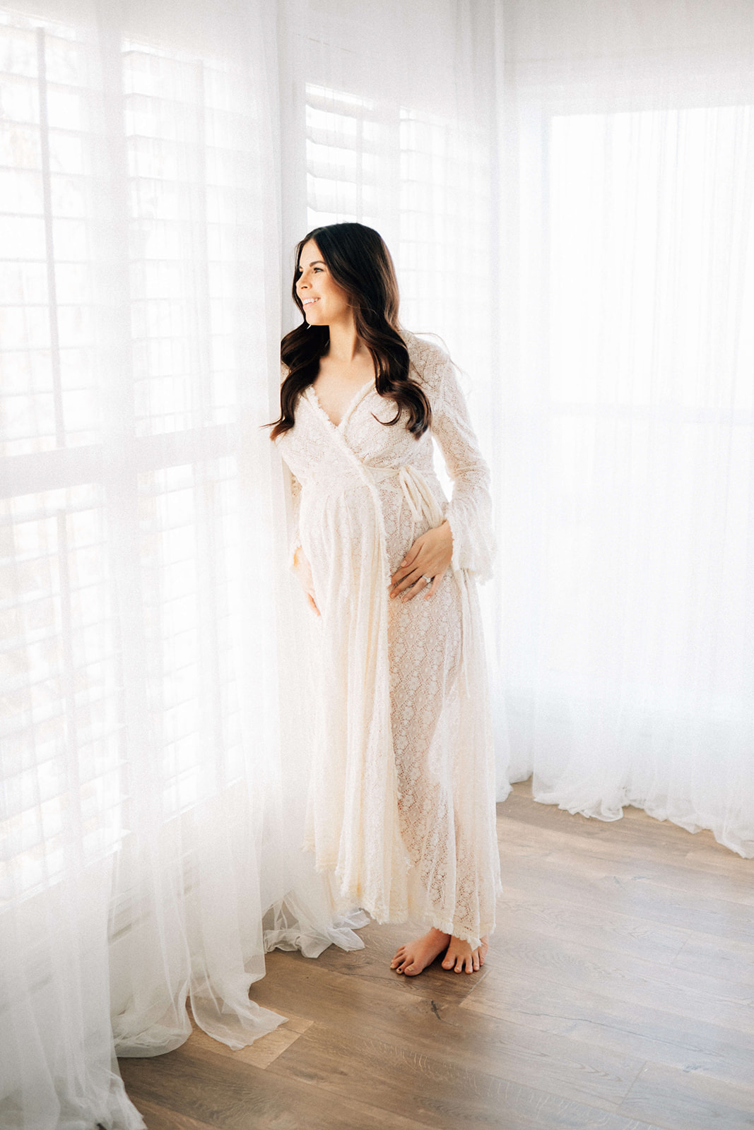 A smiling mother to be leans in a tall window wearing a lace maternity robe after some prenatal yoga in Denver