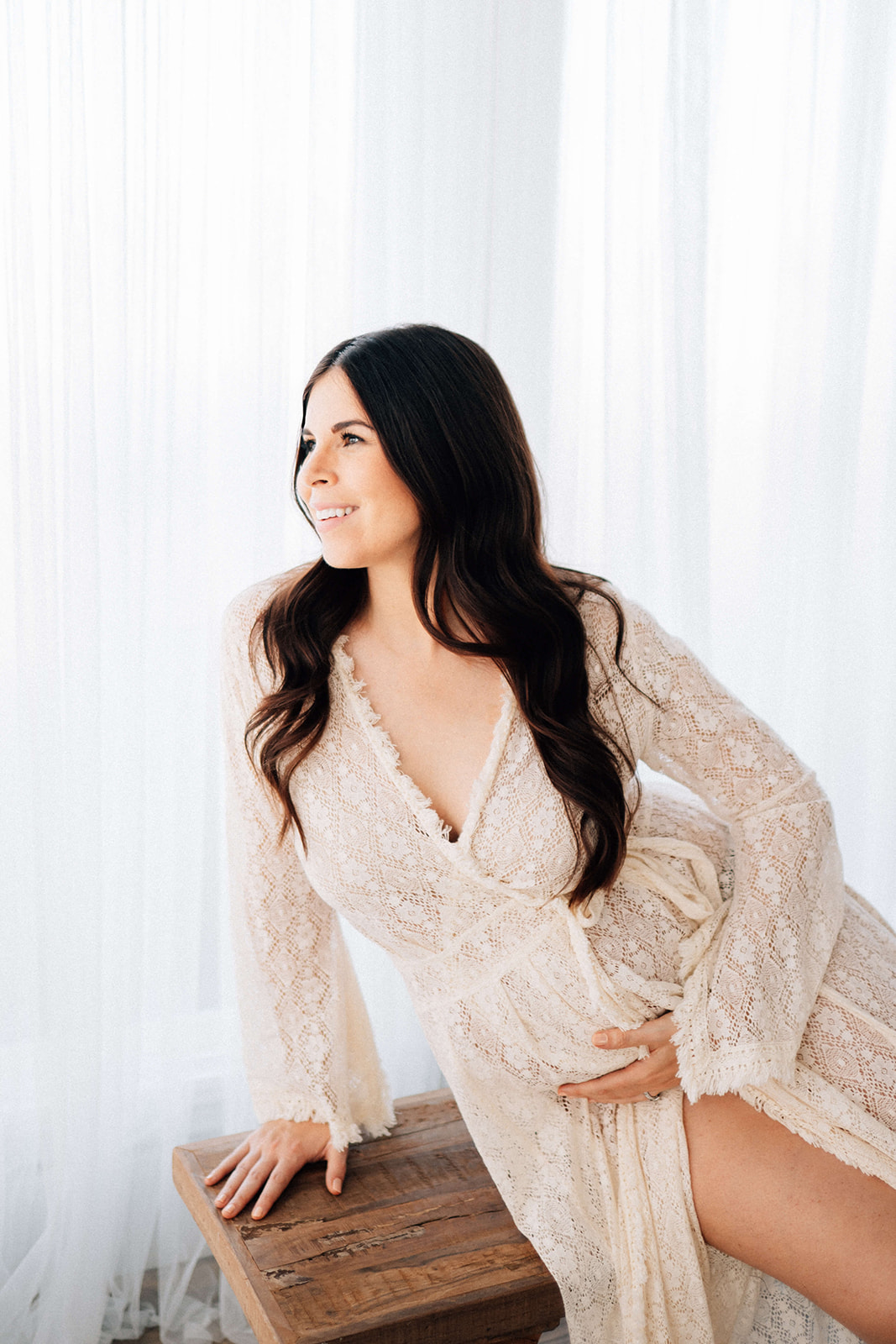 A mom to be in a lace maternity gown leans on a wooden bench smiling in a studio after some prenatal yoga in Denver
