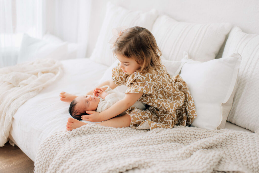 toddler girl sitting on family photo bed with her legs outstretched looking down at her baby brother