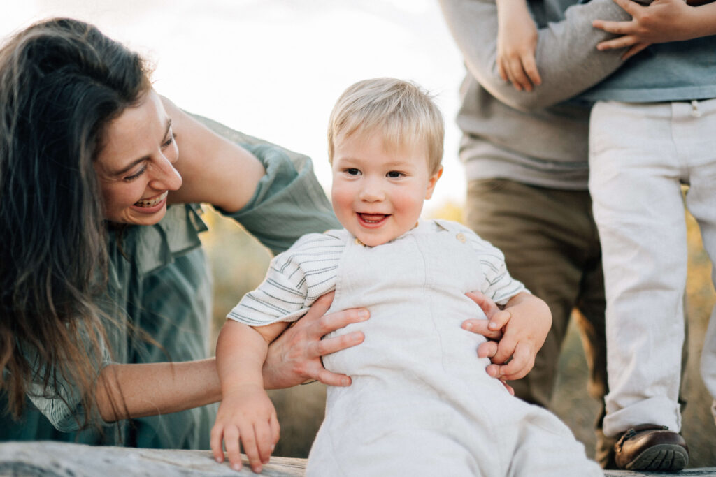 toddler boy sitting on a log with his mom holding him and his dad and brother to the side