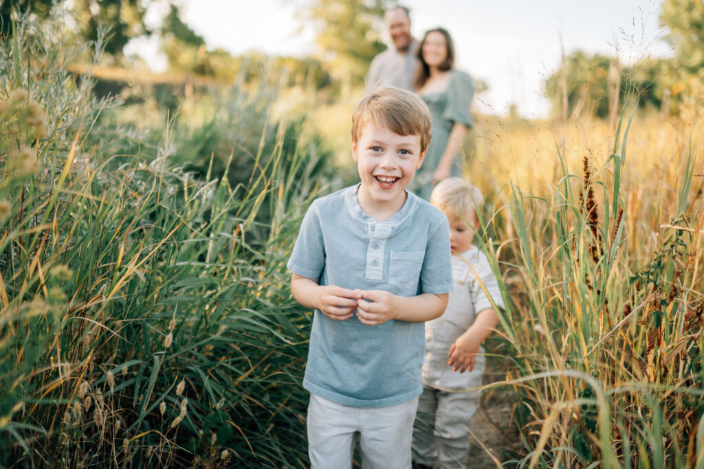 a young boy walking down a grassy path with his little brother close behind him and his parents looking on