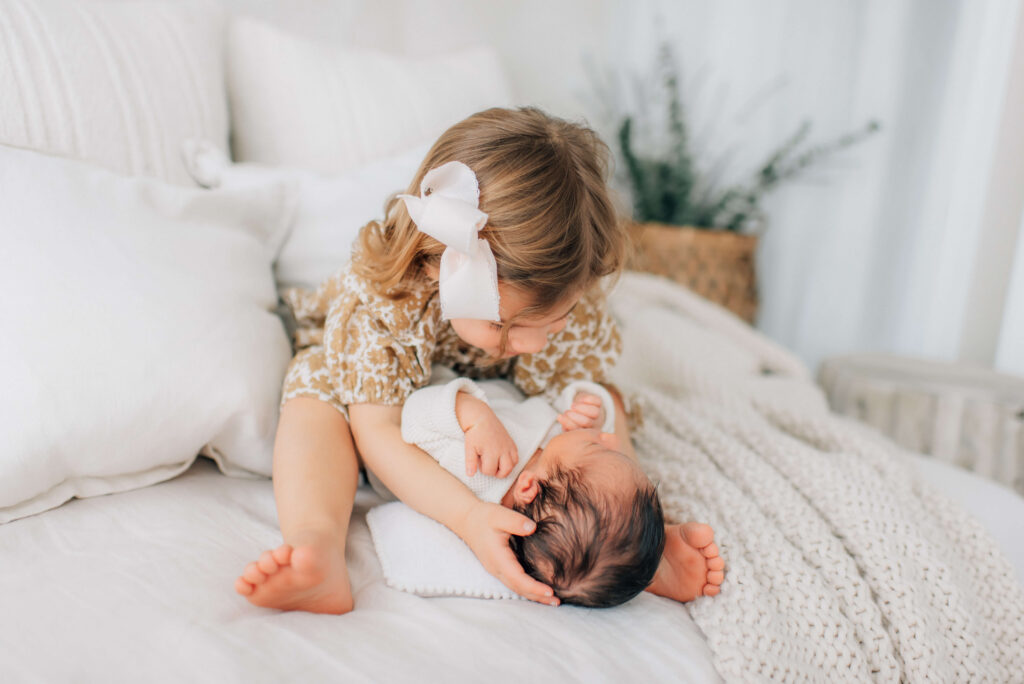 toddler sister holding her newborn brother between her legs and looking down at him