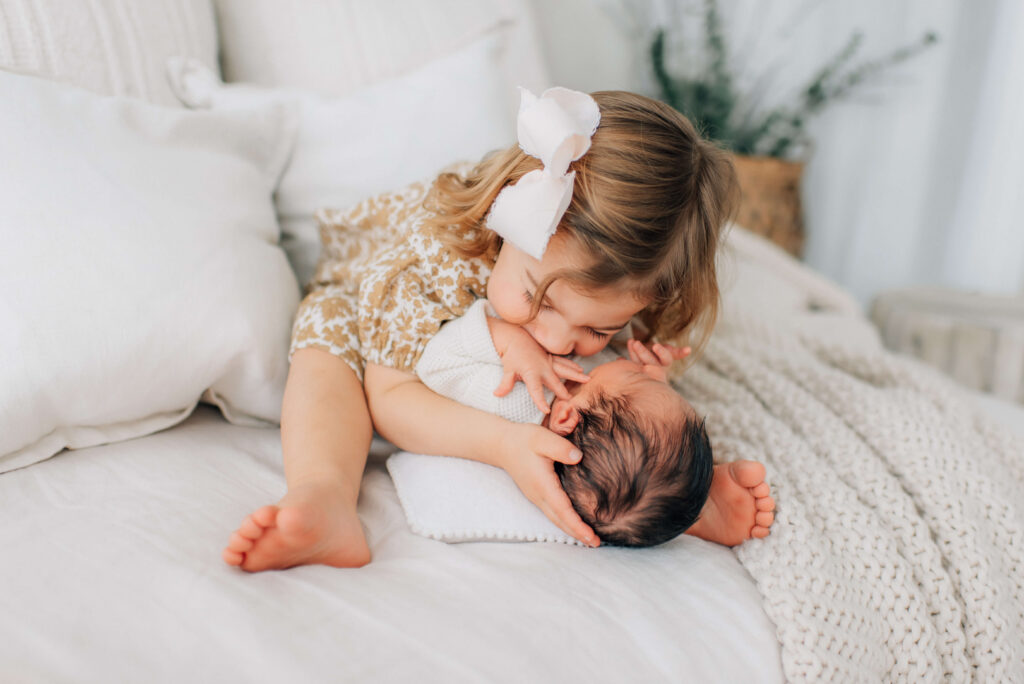 newborn baby boy lying on a bed between toddler sister's legs while she hugs him