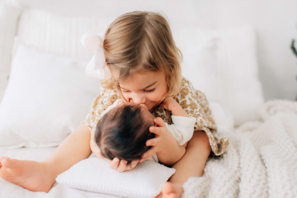 toddler sister preparing for family photos with newborn brother by holding his head and snuggling him